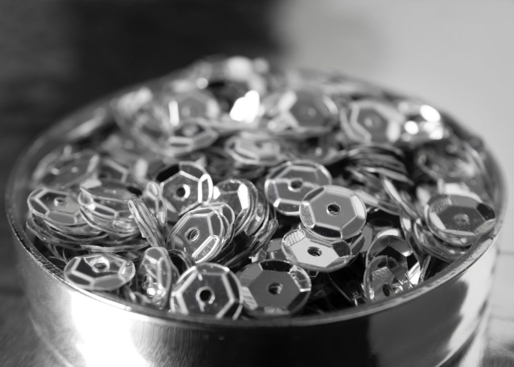 Primer plano en blanco y negro de un recipiente metálico lleno de pequeñas piezas de metal plateado facetadas con orificio central.