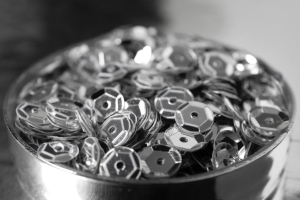 Primer plano en blanco y negro de un recipiente metálico lleno de pequeñas piezas de metal plateado facetadas con orificio central.