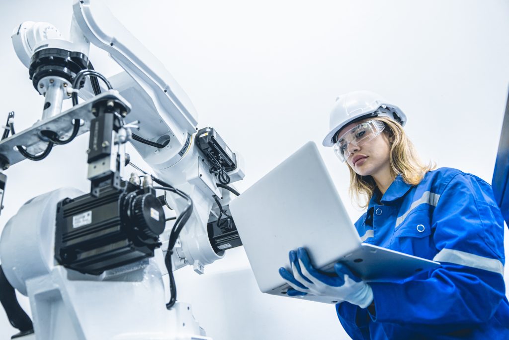 Ingeniera industrial con casco y gafas de seguridad trabajando en una laptop junto a un brazo robótico automatizado en una fábrica tecnológica.
